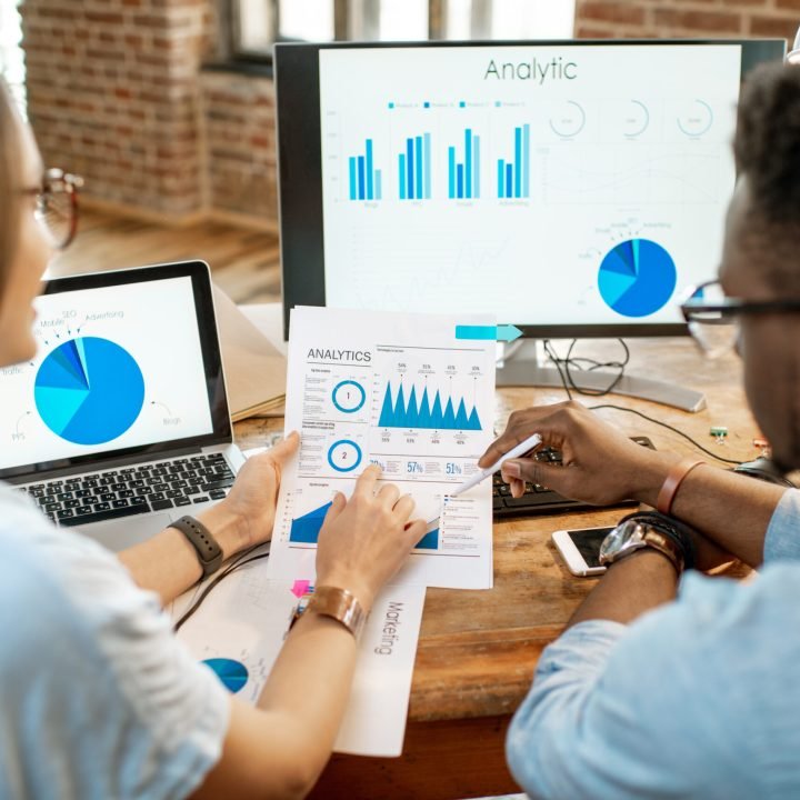 Coworkers working with analitycs sitting together at the working place with computer, laptop and documents. View from behind focused on the documents