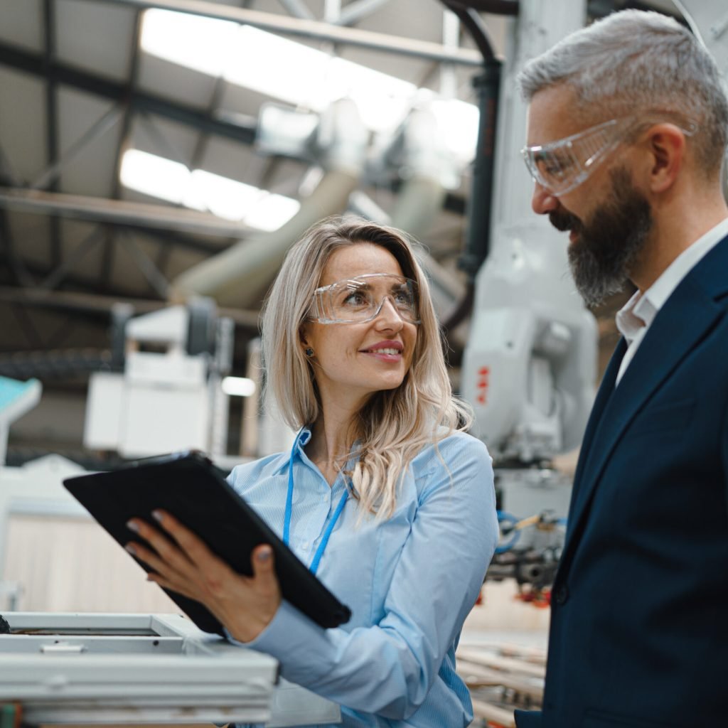Female engineer and male supervisor standing in modern industrial factory, talking about production. Manufacturing facility with robotics, robotic arms and automation.